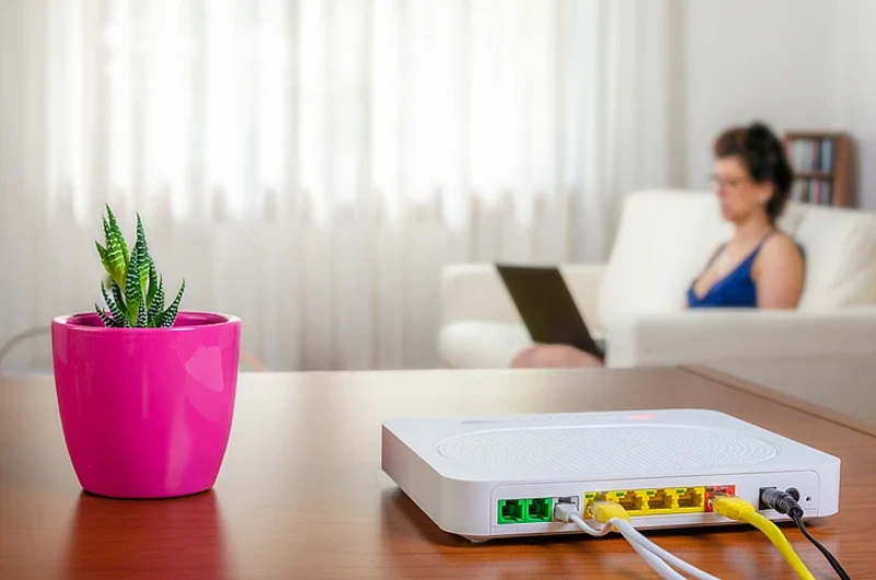 Roteador doméstico em primeiro plano e mulher usando notebook ao longe.